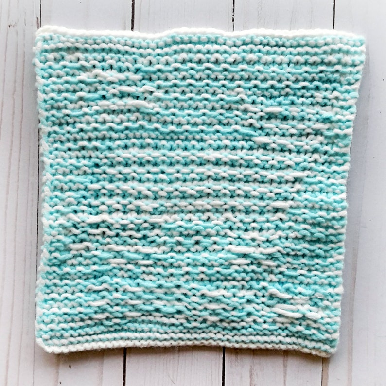 May include: A knitted dishcloth in a light blue and white colour scheme. The cloth is made with a simple knit stitch and has a slightly textured surface.