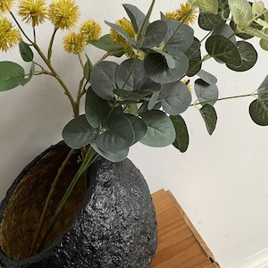 May include: A black, textured ceramic vase with a gold interior, holding a bouquet of green eucalyptus and yellow pom-pom flowers.