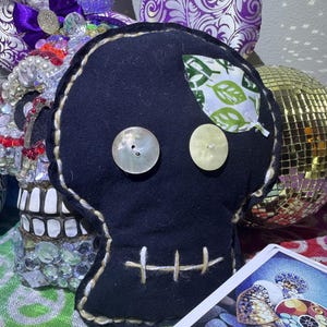 May include: A black skull-shaped plush doll with button eyes and a stitched mouth. A leaf-shaped patch with a green and white pattern is on the forehead. The doll is surrounded by decorative items, including a disco ball and tarot cards.