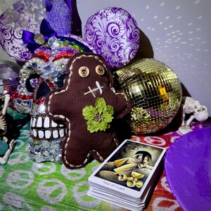 May include: A brown fabric doll with button eyes and a green flower accent, positioned in front of a stack of tarot cards. A disco ball and decorative skull are also visible. The tarot cards are labeled "Four of Cups".