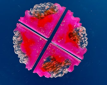 May include: Four-piece coaster set. Each coaster is a vibrant magenta with a scalloped edge and a central design of red and silver leaf accents. The coasters are arranged in a circular pattern.