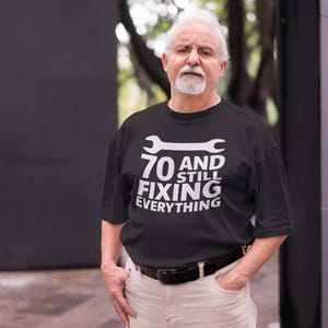 Könnte beinhalten: Schwarzes T-Shirt mit dem weißen Text "70 AND STILL FIXING EVERYTHING" und einer Schraubenschlüssel-Grafik. Das T-Shirt wird von einer Person mit weißem Bart und heller Hose getragen.