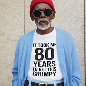 May include: An older person wearing a red beanie, sunglasses, and a light blue cardigan over a white t-shirt. The t-shirt has the text "IT TOOK ME 80 YEARS TO GET THIS GRUMPY" in black. The person is also wearing a black belt and light blue pants.