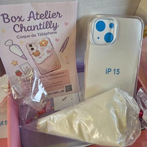 Peut inclure: Une boîte rose contenant une coque d'iPhone 15, une poche à douille remplie de matière blanche et un kit de décoration. La coque est transparente avec des accents bleus et le texte "iP 15". La boîte porte le texte "Box Atelier Chantilly".