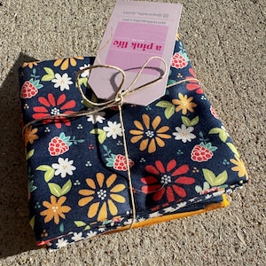 Può includere: Un fascio di tessuto piegato con un motivo floreale di fiori rossi, arancioni e bianchi e bacche rosse su sfondo blu navy. Il fascio è legato con spago e include un'etichetta che recita "a pink life quilts".