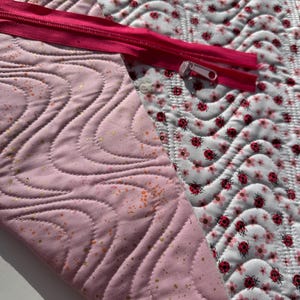 May include: A pink quilted fabric with a gold glitter pattern and a pink zipper. The fabric is sewn to a white fabric with a pattern of red ladybugs and white flowers.