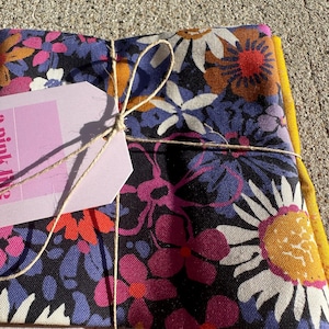 Puede incluir: Un conjunto de cuadrados de tela floral atados con hilo y una etiqueta rosa. La tela presenta un vibrante estampado de grandes flores blancas, rosas y naranjas sobre un fondo azul oscuro, con un borde amarillo. La etiqueta está en blanco.