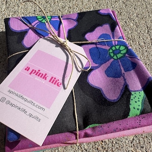 May include: A stack of fabric squares tied with twine, featuring a floral pattern in purple, blue, and green on a black background. A pink tag with the text "a pink life quilts" is attached.