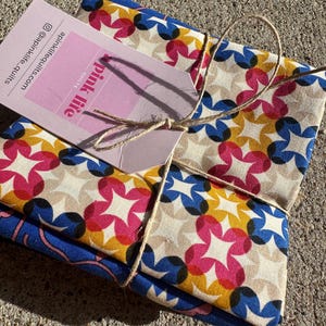May include: A stack of fabric squares tied with twine. The fabric features a repeating pattern of colorful, overlapping shapes in pink, blue, yellow, and tan. A pink tag with the words "Pink Life Quilts" is attached.