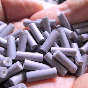 May include: A collection of small, cylindrical beads in a muted lavender-gray color. Each bead features a smooth, matte finish and a central hole running through its length, revealing a white interior core visible at the ends of the cylinders.