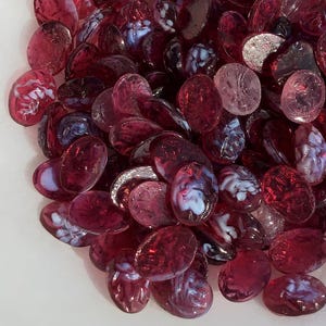 May include: A pile of oval glass beads in shades of red, burgundy, and pink. Some beads have white and blue swirls, while others are translucent. The beads are scattered, creating a textured appearance.