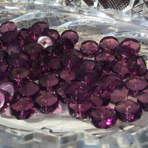 May include: A collection of faceted, deep purple beads fills a clear, cut-glass bowl. The beads have a multi-faceted design, catching the light and reflecting a range of purple hues. The bowl's intricate cut-glass pattern adds to the overall aesthetic. These beads are suitable for jewelry making.