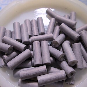 May include: A collection of cylindrical beads in a muted lavender-gray color with a subtle mottled texture. Each bead features a central hole running through its length. The beads are arranged in a pile within a circular, off-white container.