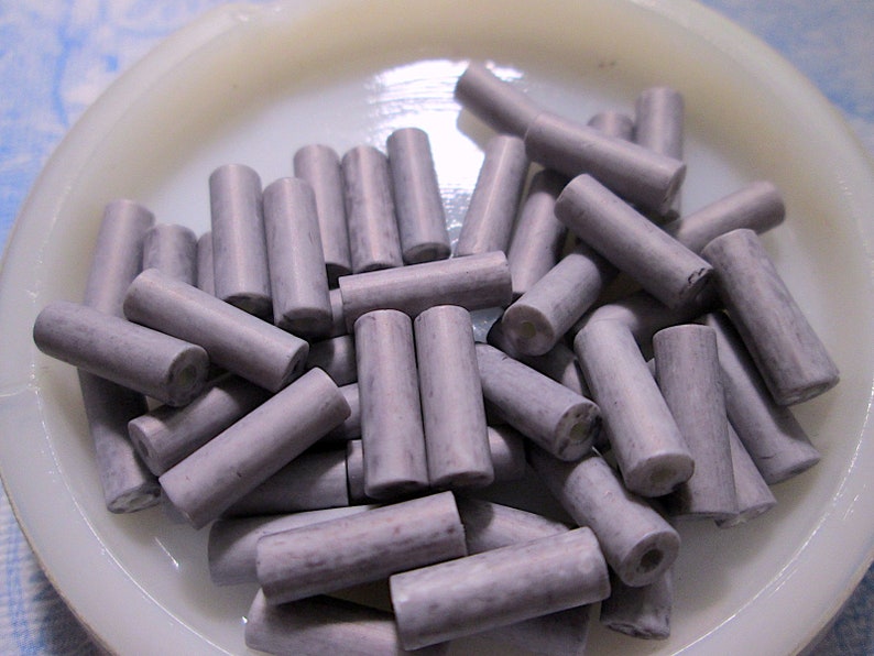 May include: A collection of cylindrical beads in a muted lavender-gray color with a subtle mottled texture. Each bead features a central hole running through its length. The beads are arranged in a pile within a circular, off-white container.