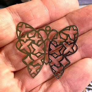 May include: A gold-colored, openwork butterfly pendant with a delicate design. The butterfly's wings feature a pattern of intersecting shapes. The pendant has a small loop at the top for hanging.