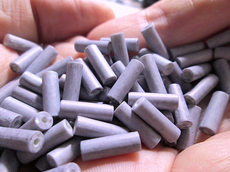 May include: A collection of small, cylindrical beads in a muted lavender-gray color. Each bead features a smooth, matte finish and a central hole running through its length, revealing a white interior core visible at the ends of the cylinders.