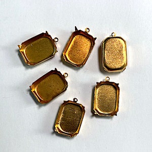 May include: Six gold-toned metal pendant trays with a rectangular shape and a beveled edge. The trays are designed to hold small items, such as stones or charms.