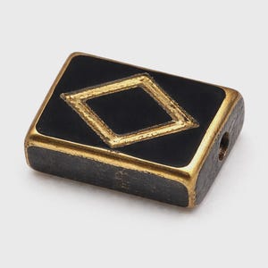 Puede incluir: Una cuenta rectangular negra con un diseño dorado en forma de diamante en el centro. Los bordes de la cuenta también son dorados. Tiene un agujero perforado en un extremo, lo que sugiere que es para ensartar o hacer joyas.