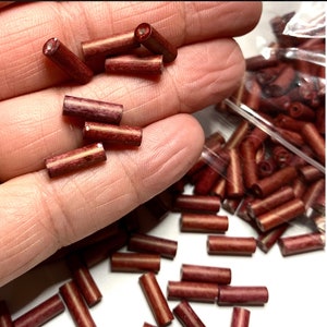May include: A bag of small, red wooden beads. The beads are cylindrical in shape and have a light brown stripe running down the centre. The beads are approximately 1.25 cm long.