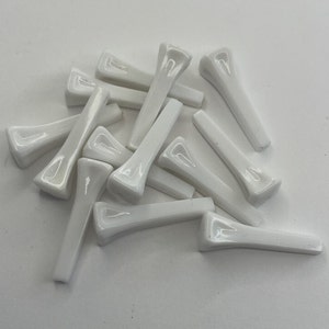 Puede incluir: Una colección de doce tees de golf blancos dispuestos en un montón disperso. Cada tee presenta un acabado liso y brillante con una cabeza ancha y cóncava y un eje cónico, diseñado para sostener una pelota de golf en el área de salida.