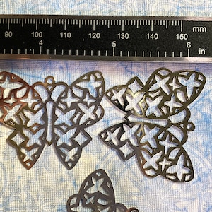 May include: Several bronze-colored metal butterfly charms with intricate cut-out designs. A ruler with inch and millimeter measurements is in the background. The butterflies are approximately 1.5 inches wide.
