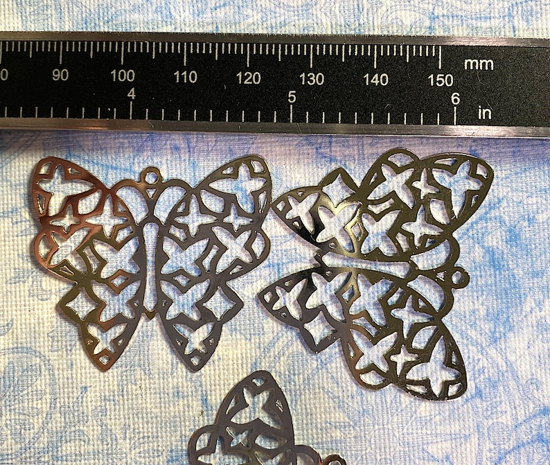 May include: Several bronze-colored metal butterfly charms with intricate cut-out designs. A ruler with inch and millimeter measurements is in the background. The butterflies are approximately 1.5 inches wide.
