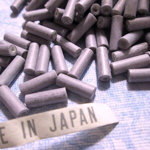 May include: A collection of small, cylindrical, light purple beads with a speckled texture and a central hole running through each piece. A white fabric tag with the black printed text MADE IN JAPAN is positioned in the foreground of the beads.