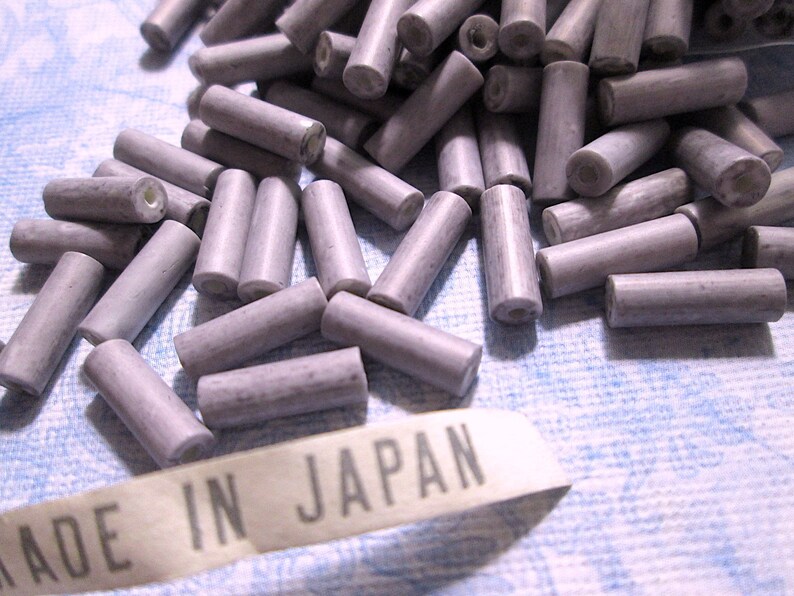 May include: A collection of small, cylindrical, light purple beads with a speckled texture and a central hole running through each piece. A white fabric tag with the black printed text MADE IN JAPAN is positioned in the foreground of the beads.