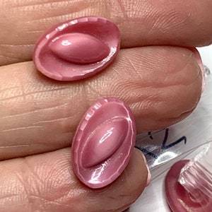 May include: Close-up of two oval-shaped, pink beads. Each bead has a raised, oval center and a textured, ridged border. The beads are a soft, muted pink color, and appear to be made of a smooth, glossy material. The beads are held in a hand.