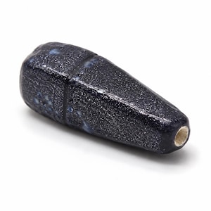 May include: A tapered, elongated ceramic bead with a dark navy blue, speckled glaze finish. The surface features a subtle, darker horizontal band near the rounded end, with small, light blue circular accents and a visible central hole at the tip.