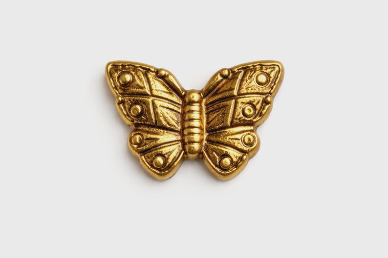 May include: A detailed, golden butterfly ornament with intricate patterns on its wings and body. The butterfly is centered on a white background, showcasing its metallic sheen and textured surface. This decorative piece could be used for jewelry or crafts.