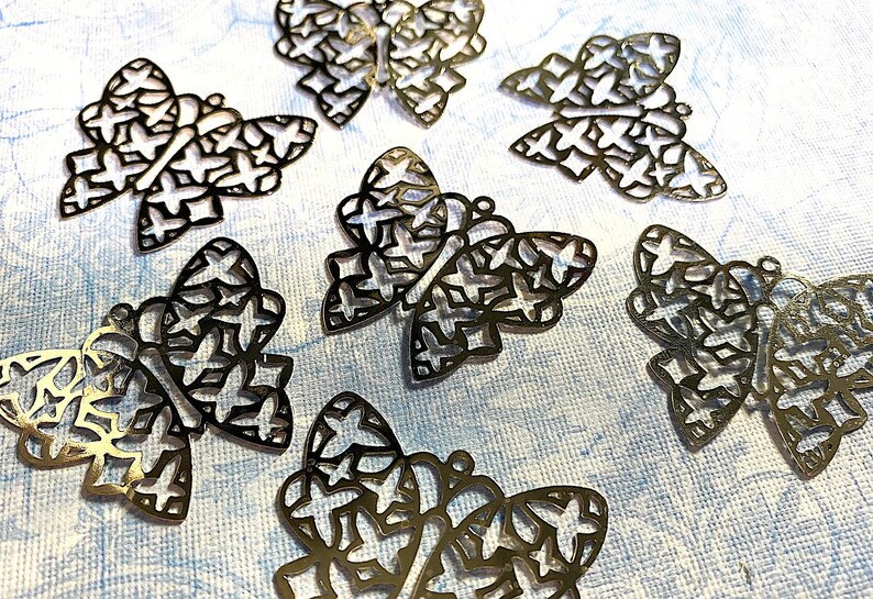May include: Close-up of several gold-colored butterfly charms. Each charm features a detailed openwork design with a butterfly shape and star-shaped cutouts. The charms are scattered on a light blue textured surface.