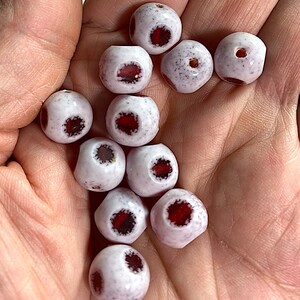 Puede incluir: Una colecci&oacute;n de cuentas de vidrio blanco con acentos rojos y negros. Las cuentas tienen una textura moteada y un agujero en el centro.