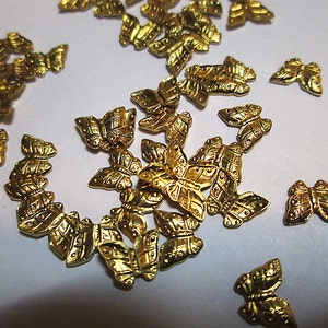 May include: A collection of small, gold-colored butterfly embellishments. Each butterfly has detailed wing patterns and a textured surface. These decorative elements are scattered across a white surface, suggesting their use in crafts or jewelry making.