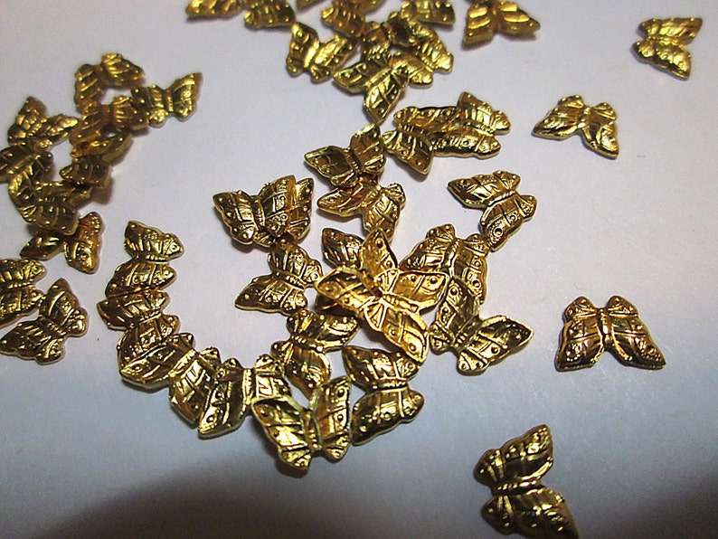 May include: A collection of small, gold-colored butterfly embellishments. Each butterfly has detailed wing patterns and a textured surface. These decorative elements are scattered across a white surface, suggesting their use in crafts or jewelry making.