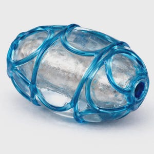 May include: An oval-shaped, clear glass object with a silver interior, encased in a vibrant blue, cage-like structure. The blue structure is made of curved lines that wrap around the glass, creating a decorative design. The object is set against a plain white background.