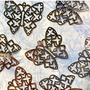 May include: A collection of metal butterfly charms with intricate star-shaped cutouts. The charms are in various shades of bronze and silver, scattered on a light blue patterned surface. Each butterfly has a small loop for attaching to jewelry or crafts.