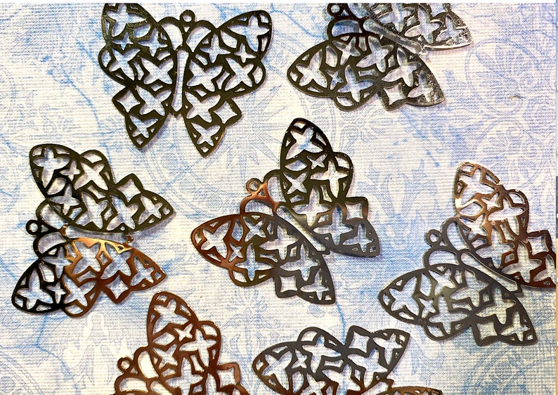 May include: A collection of metal butterfly charms with intricate star-shaped cutouts. The charms are in various shades of bronze and silver, scattered on a light blue patterned surface. Each butterfly has a small loop for attaching to jewelry or crafts.