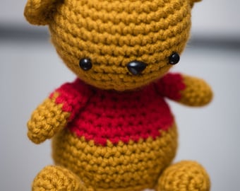 Op de afbeelding: Een handgemaakte gehaakte teddybeer in goud- en roodtinten. De beer heeft een ronde kop, kleine zwarte ogen en een zwarte neus. Hij draagt een rood shirt en heeft afgeronde ledematen. Het speelgoed staat op een reflecterend oppervlak. Ongeveer 15 cm hoog.
