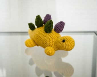 May include: A handmade crochet dinosaur toy in a vibrant yellow. The stegosaurus features green and purple dorsal plates, small yellow legs, and a black eye. This soft toy is a charming addition to any child's collection.