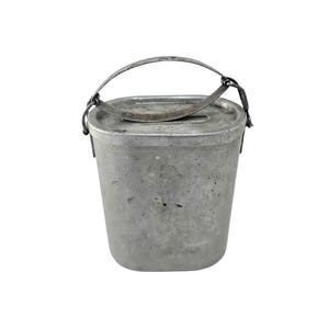 Puede incluir: Un recipiente metálico rectangular vintage con tapa abisagrada y asa curva. El recipiente es de color plateado apagado y parece estar hecho de aluminio. El asa está sujeta a los lados con bisagras metálicas.