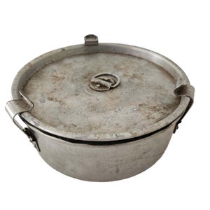 May include: A round, silver-colored metal container with a lid. The lid has a small handle and is secured by metal clips. The container appears to be vintage or antique, with signs of wear and age.