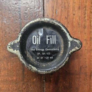 Può includere: Un tappo di riempimento dell'olio nero vintage con scritte bianche. Il testo sul tappo recita "Oil Fill" e "Use Energy Conserving". Il tappo ha un aspetto usurato e invecchiato.