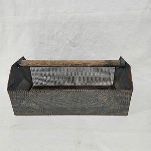 May include: A rectangular metal tool caddy with a perforated design and a wooden handle. The caddy is a dark gray color and has a rustic, vintage appearance. The handle is made of light brown wood.