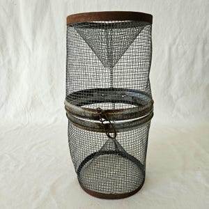 Könnte beinhalten: Eine antike Fischfalle aus Metall mit zylindrischer Form. Die Falle besteht aus Drahtgeflecht und hat einen rostigen Metallrand oben und unten. Ein scharnierter Metallverschluss sichert den Mittelteil. Innen konisch. Maße in cm.