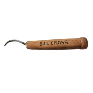 Może przedstawiać: Nóż do rzeźbienia w drewnie z zakrzywionym ostrzem. Drewniany uchwyt jest cylindryczny i ma nadruk "BILL CROSS". Ostrze jest metalowe i zakrzywione.