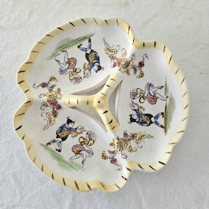 Pode incluir: Uma travessa de servir vintage de três secções com borda ondulada. A travessa de cerâmica branca suja apresenta figuras pintadas à mão em roupas coloridas, com uma borda amarela e uma pega central. A travessa tem aproximadamente 25 cm de diâmetro.