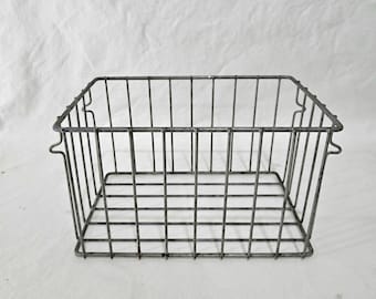 May include: A rectangular, open-top wire basket with a distressed, gray finish. The basket features vertical and horizontal wire bars, with small handles on the sides. The basket is designed for storage and organization.