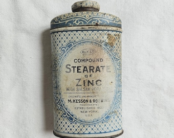 May include: Vintage cylindrical tin of McKesson & Robbins Compound Stearate of Zinc with Balsam Peru. The tin has a blue and white patterned design with text that reads "McKesson & Robbins, Incorporated, Established 1833, New York, U.S.A."