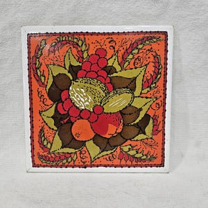 May include: Square ceramic tile with a vibrant fruit design. The artwork features red grapes, oranges, and nuts, surrounded by green leaves and wheat stalks, set against an orange background. The tile has a white border.